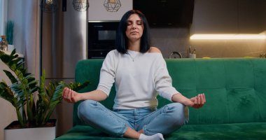 Evde meditasyon yapan çekici bir kadın, kanepeye oturup yoga ve meditasyon yaparken stresi gözleri kapalı olarak hafifletir..