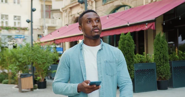 stock image Portrait of good-looking smiling happy young black-skinned guy in casual clothes which walking near street cafe and using his mobile