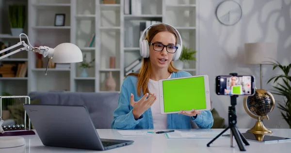 若いかなりの女性は 机の上に座って ビデオ通話会議のスピーチを行い 三脚のレコードビデオブログにスマートフォンを使用して緑の画面のデジタルタブレットを示しています — ストック写真