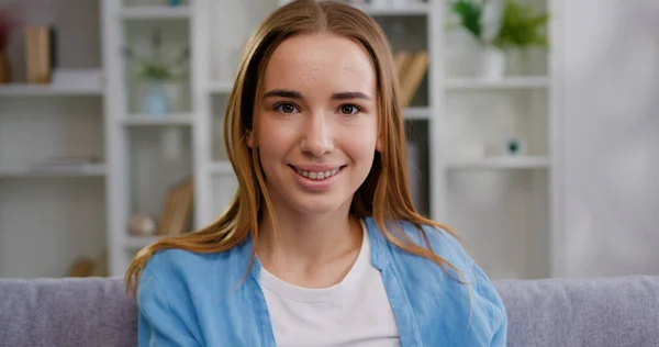 Çekici, mutlu, genç bir kadın evdeki rahat koltukta oturuyor ve gülümseyen tatmin olmuş yüz ifadesiyle kameraya bakıyor, kadın portresi.