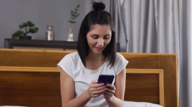 Çağdaş yatak odasında yatağında otururken telefondaki mesajları okuyan, iyi huylu, koyu renk saçlı bir kadının ön görüntüsü.