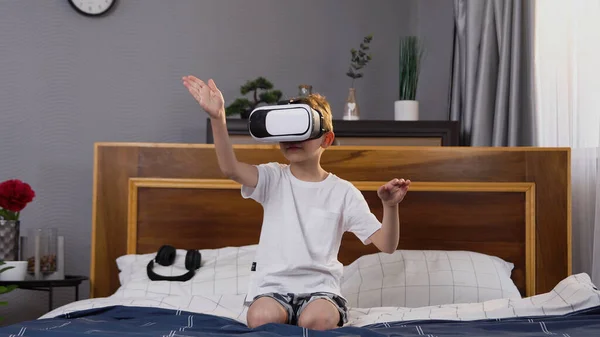 見栄えの良いコンテンツ8 10歳の男の子がベッドルームで快適なベッドに座って 拡張現実ゴーグルを適用する仮想画面上で手を動かす — ストック写真