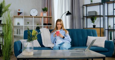 Oldukça tatmin olmuş 20 yaşında, uzun açık renk saçlı, gündelik kıyafetlerle oturma odasında rahat bir koltukta oturuyor ve akıllı telefonunu uyguluyor.