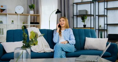 Çekici, gülümseyen, neşeli, açık renk saçlı, gündelik elbiseli bir kız evdeki koltukta oturuyor ve olumlu bir telefon konuşması yapıyor.