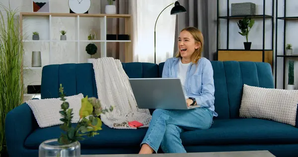 Good-looking joyful happy contented 20-aged light haired girl in casual wear sitting on comfortable sofa in contemporary apartment and revisioning funny program on computer,front view