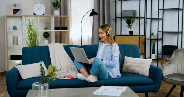 stock image Distant learning concept where good-looking smart cheerful light haired girl in casual clothes using headphones while has online video chat with teacher sitting in relaxed pose on soft sofa in