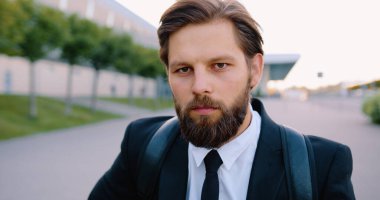 Kendine güveni tam, sakallı, sırt çantalı genç bir adamın portresi. Sokakta, büyük şehir binasının yanında poz veriyor.