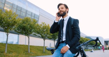 Gülümseyen, kendine güvenen, şık giysiler içinde sakallı bir adam motosikletinin yanında duruyor ve modern şehir binasının arka planında telefonla konuşuyor.