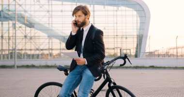 Yakışıklı, yoğun sakallı, resmi giysiler içinde bisikletine yaslanmış, cep telefonuyla konuşan ve modern şehir inşaatının yanındaki mali belgeleri okuyan genç bir iş adamı.