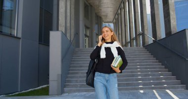 Güzel, gülümseyen, açık renk saçlı, sıradan giyinmiş bir kız öğrenci üniversiteden çıkarken merdivenlerden iniyor ve telefon konuşmasının keyfini çıkarıyor.