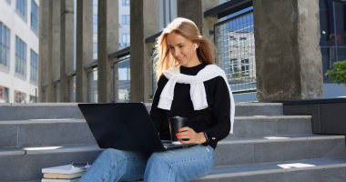 Şehir binasının basamaklarında oturan, dizüstü bilgisayarda çalışan ve kağıt bardaktan kahve içen gündelik kıyafetlerle muhteşem gülümseyen modern genç bayana yakın durun.