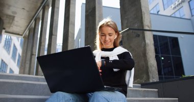 Güzel görünümlü, sakin, gülümseyen, 20 yaşında, açık renk saçlı bir kızın portresi. Binaların merdivenlerinde oturuyor, dizüstü bilgisayarda çalışıyor ve el saatini kontrol ediyor.