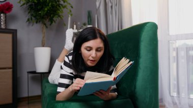 Evdeki yumuşak kanepede uzanıp kitap okuyan, gamsız, gamsız, siyah saçlı genç bir kadını yakından çek.