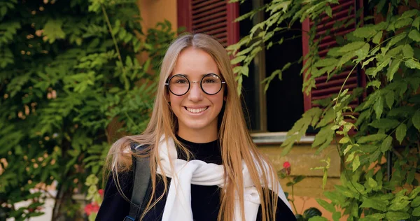 Gorgeous smiling happy light-haired girl in stylish glasses standing in front of camera near ancient city building with green plants ,close up