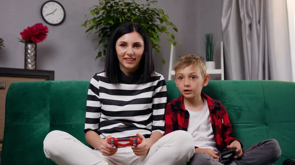 ゲームパッドを使用して設定されたテレビでビデオゲームを楽しむ母親と息子の肯定的な感情的な幸せな家族の肖像画 自宅で快適なソファに座って — ストック写真
