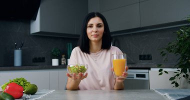 Güzel dekore edilmiş bir mutfakta kameraya poz veren pozitif görünüşlü mutlu esmer ve cam kasede taze hazırlanmış sebze salatası ve cam bardakta lezzetli portakal suyu.