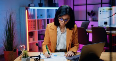 Resmi giyim ve gözlük takan oldukça kendine güvenen esmer, akşam ofisinde bilgisayar ve finansal kağıt raporlarıyla çalışan, iş konsepti