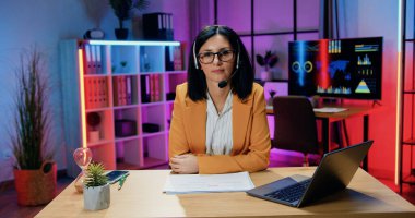 Oldukça kendine güvenen, kulaklıklı, şık giyinen, gözlüklü, laptopta çalışan ve akşam ofisinde dostça bir yüz ifadesiyle kameraya bakan modern, deneyimli bir esmer.