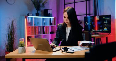 Güzel, kendine güvenen, çalışkan, koyu renk saçlı iş kadını dizüstü bilgisayarda çalışırken giyiniyor ve akşam ofisindeki önemli verileri not ediyor.