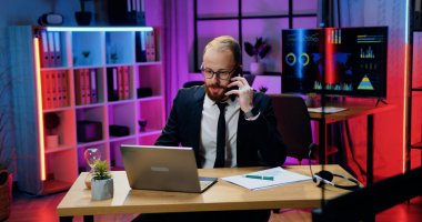 Ofiste çalışan yakışıklı, yetenekli, yetenekli, sakallı iş adamı resmi gözlüklü, telefonla konuşan, kağıt raporlarını ve bilgisayar bilgilerini inceleyen.