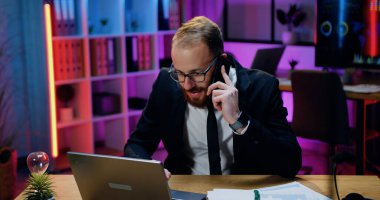 Çekici olumlu deneyimli başarılı sakallı iş adamı iş arkadaşı ya da müşterisiyle telefonda konuşuyor ve akşam ofisindeki laptop ekranına bakıyor.