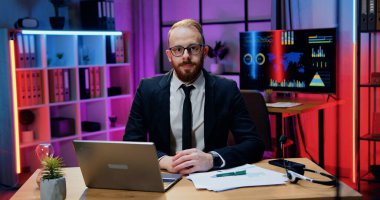 Kendine güveni tam, profesyonel, sakallı, resmi gözlüklü bir iş adamı akşam ofisinde kameranın önünde oturuyor ve dostça bir yüz ifadesiyle bakıyor.