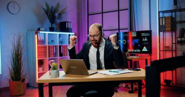 İş başarısı konsepti, resmi giyinmiş ve kulaklık takmış yakışıklı, neşeli, çalışkan iş adamının akşam çalışma odasında dizüstü bilgisayarında iyi haberlerin keyfini çıkardığı yer.