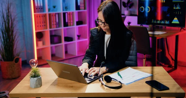 Çekici, ciddi, deneyimli, koyu renk saçlı iş kadınının portresi. İş yerinde oturan ve geç saatlerde dizüstü bilgisayarda yazan siyah elbiseli iş kadını.