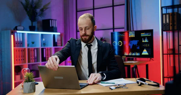 Handsome concentrated industrious responsible skilled bearded businessman sitting at his workplace in evening office and working on computer