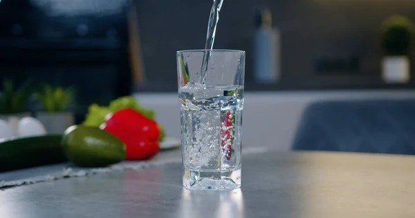 Sebzelerin yanında masanın üstünde duran şeffaf cam, çağdaş mutfak arka planında taze suyla doludur.