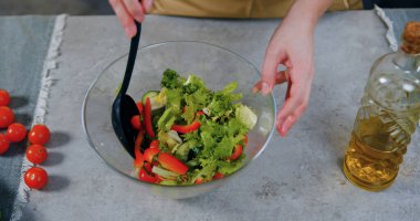 Taze sebze salatasını karıştıran şeffaf kâseye kaşık süren ve ailesi için besleyici yemek hazırlayan kadın ellerinin üst görüntüsü.