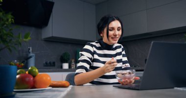 İyi görünümlü, rahat ve rahat genç bir kadın laptopta ilginç videolar izliyor ve yoğurtla mısır gevreği yiyor.