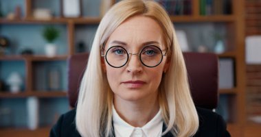 İyi donanımlı bir ofiste gözlüğünü çıkarırken kameraya bakan başarılı yetişkin kadın ofis müdürünün iş portresi.