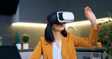 Sevimli akıllı 35 yaşında başarılı stil sahibi iş kadını mutfak masasında oturuyor ve arttırılmış gerçeklik kulaklığıyla sanal ekran üzerinde çalışıyor.