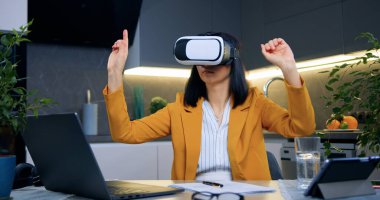 Güzel, kendine güvenen, şık giysiler içinde başarılı bir iş kadını masanın yanındaki çağdaş mutfakta oturuyor ve sanal gerçeklik kulaklığı takıyor.