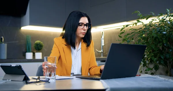 stock image Front view of attractive successful hardworking adult brunette in stylish clothes which drinking fresh water from glass while remotely working on laptop at home office