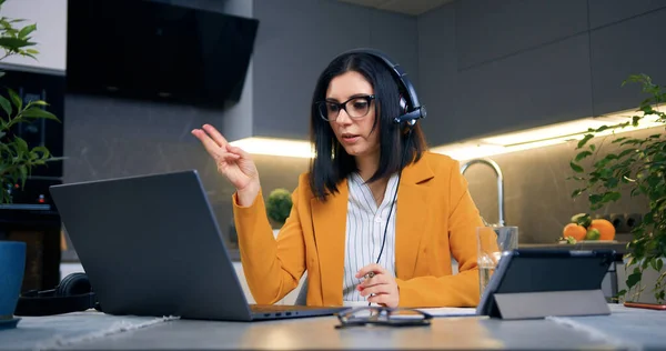 Beautiful experienced confident adult black-haired woman in glasses and stylish jacket sitting in front of laptop in headset and holding online meeting with client or worker from home