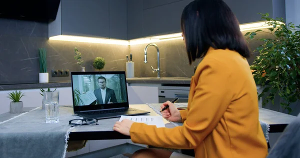 Beautiful attantive experienced black-haired woman in orange jacket sitting in front of computer in contemporary kitchen and listening explaining from business partner via video conference