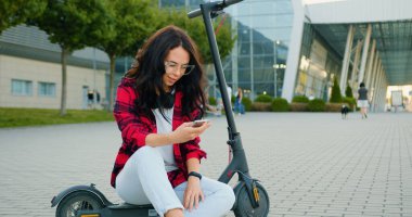 Hoş, olumlu, modern stil sahibi, uzun kahverengi saçlı, boynunda gözlük ve kulaklık takan, kendi e-scooter 'ında oturan ve cep telefonu kullanan genç bir kadının portresi.