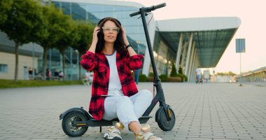 Çekici, pozitif, güler yüzlü, günlük giysiler içinde, kişisel e-scooter 'a binmiş, kulaklık takmış, şehir caddesinde, ön tarafta yürürken en sevdiği müziği dinleyen genç bir kadın.