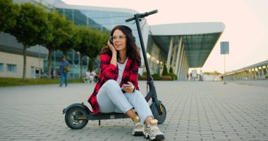 Güzel, gülümseyen, tasasız, genç esmer, gözlüklü ve modaya uygun giysileri olan şehirli modern mobiletin yanında e-scooter 'da otururken telefon çalma listesindeki şarkıların tadını çıkaran genç bir kızın ön görüntüsü.