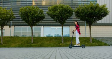 Büyük şehir binasının yanında yeşil bitkiler ve ekolojik e-scooterla gezen sıradan giysiler içindeki çekici, mutlu, genç esmer kız.