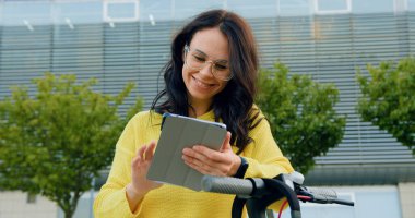 Sevimli arkadaş canlısı, güler yüzlü, şık giyinmiş, gözlüklü, tablet bilgisayarını kullanan esmer bir kız. Gündüz vakti büyük modern bina arka planında elektrikli scooterının yanında duruyor.