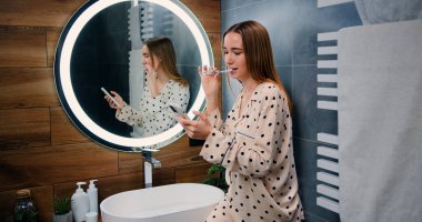 Young happy smiling woman is using smartphone for work or entertainment while brushing her teeth with toothpaste in bathroom at home.