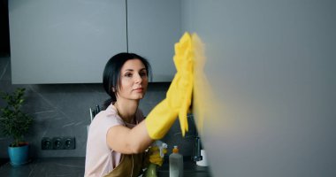 Ev mutfağındaki mutfak yüzeyini temizlerken önlüklü ve koruyucu eldivenli ciddi beyaz kadın.