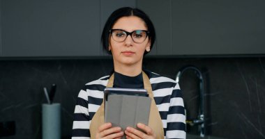 Çekici, yoğun, mutfak önlüklü, elinde tablet olan, kendi mutfağında çalışan kadının portresini kapat.
