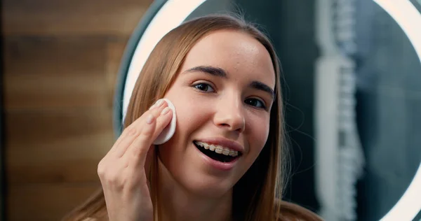 Bornozlu güzel sarışın kadın kameraya bakıp pamuk pedleri ve temiz bir cilt kullanıyor. Cilt bakımı, profesyonel bakım ürünü kozmetik reklam kavramı.