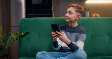 9 yaşında güzel bir beyaz çocuk kanepede oturuyor ve akıllı telefonuyla oyun oynuyor. Cep telefonuyla oynayan bir genç. Çocuk oyun için telefon kullanıyor..