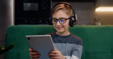 Evde kulaklıkla dijital tablet tarama, canlı yayın ve video izleme kullanan sevimli, güler yüzlü çocuğu kapatın. Evdeki kanepede oturan tablet bilgisayarlı güzel genç adam.