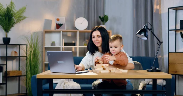 Güzel görünüşlü mutlu esmer iş yerinde dizüstü bilgisayarda çalışıyor ve dizlerinin üzerinde küçük şirin oğlu tahta bloklarla oynuyor.